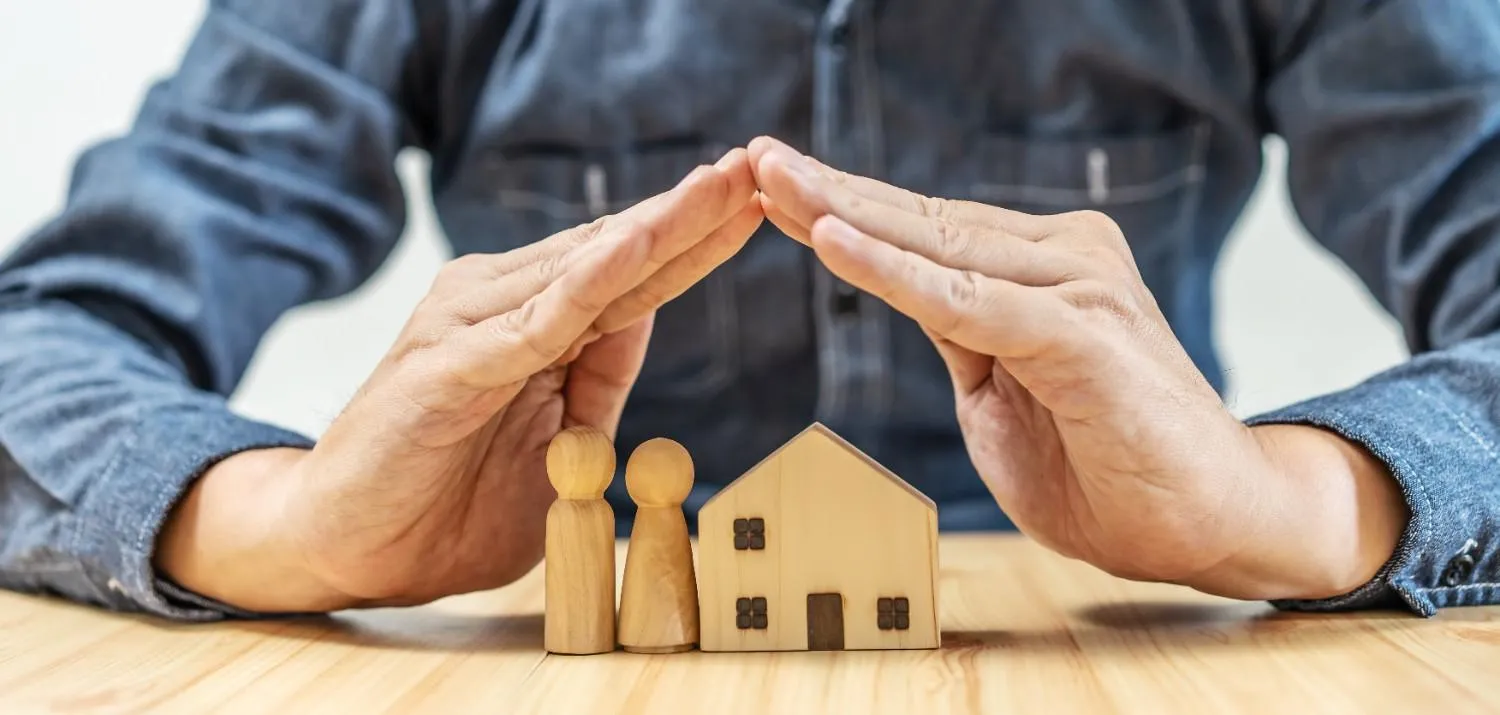 Hands forming a roof over a wooden house and two small figures